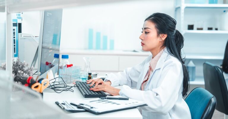 Una scienziata in camice da laboratorio digita su una tastiera mentre analizza i dati di una ricerca al computer. Sono visibili attrezzature di laboratorio e campioni chimici, a dimostrazione di un ambiente di lavoro scientifico e concentrato