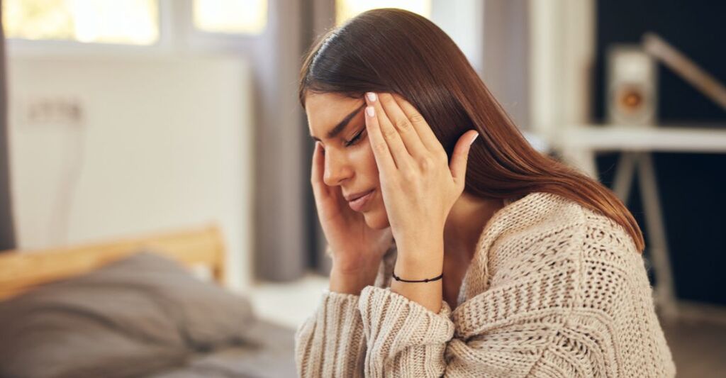 Attraente bruna caucasica vestita con un maglione beige seduta sul letto in camera da letto e con mal di testa.