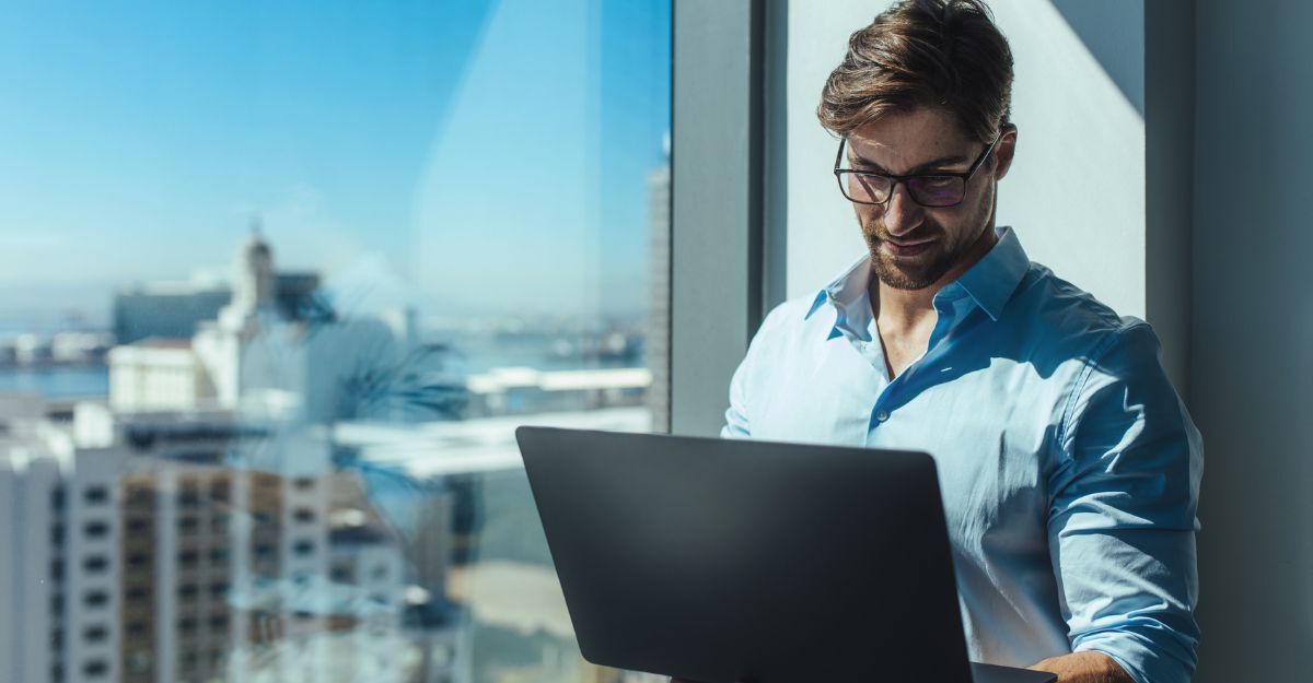Uomo con occhiali lavora al portatile in piedi vicino a una finestra in un ufficio con vista sulla città.