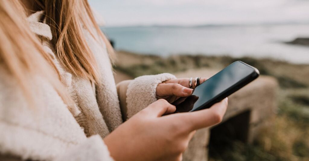 Primo piano delle mani che tengono uno smartphone all'aperto con uno sfondo marino sfocato.