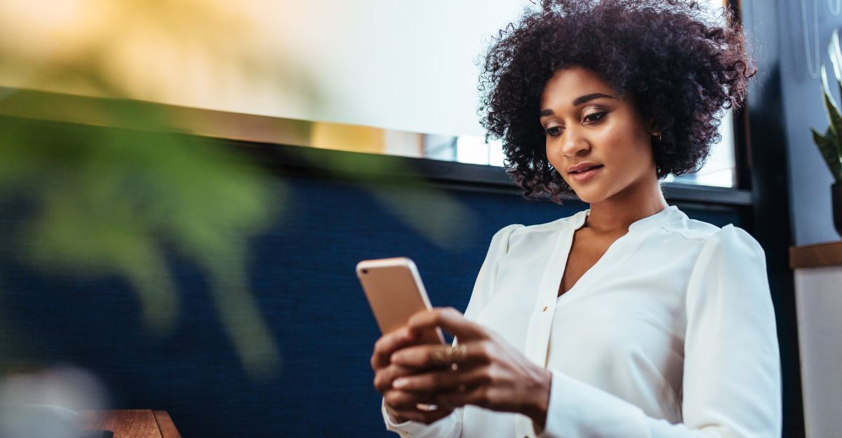 Donna con capelli ricci che guarda il suo smartphone in un ambiente interno.