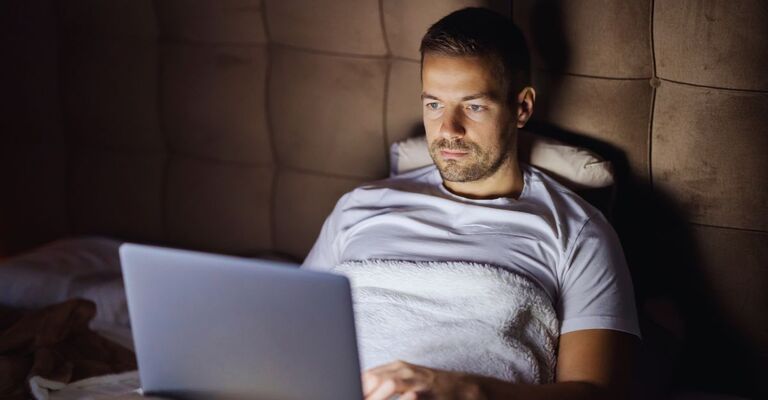 Un giovane uomo sta usando il suo computer portatile a letto, a tarda notte, in una camera da letto buia