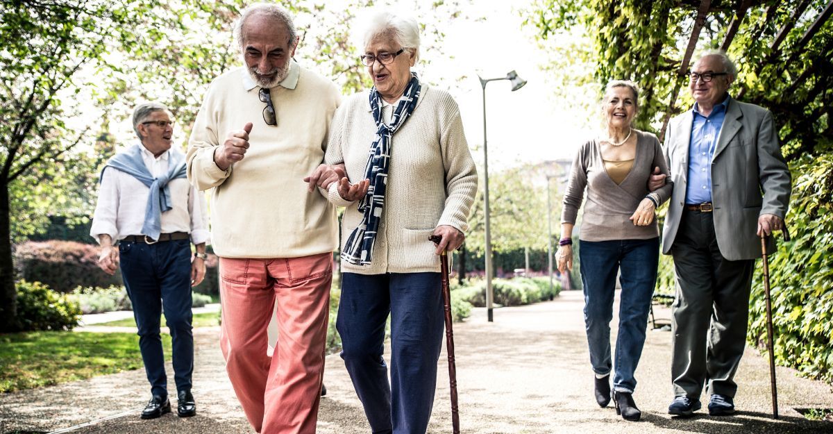 Cinque persone anziane camminano insieme in un parco alberato. Due coppie in primo piano si tengono a braccetto o si sostengono, mentre un uomo cammina da solo sullo sfondo a sinistra.
