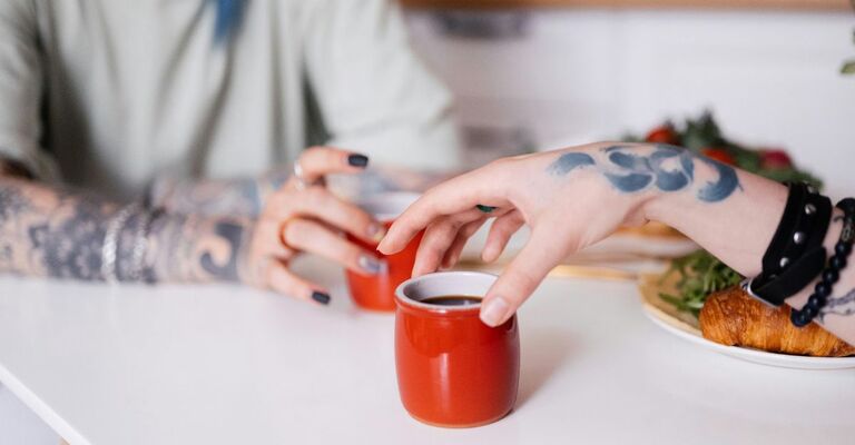 Primo piano di due persone con le mani tatuate che prendono insieme il caffè in delle tazzine rosse sopra il tavolo della cucina bianco