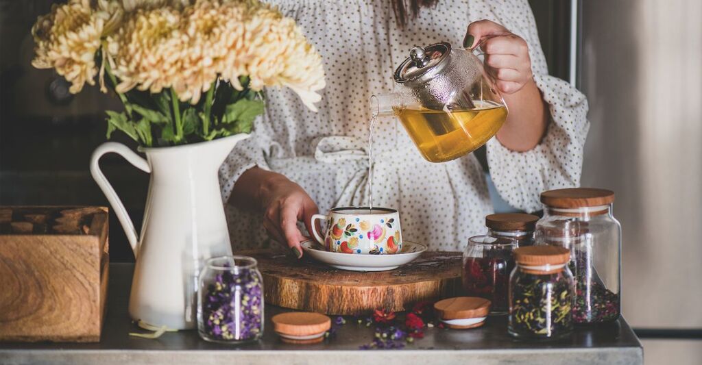 Giovane donna caucasica che versa il tè verde appena preparato da una teiera di vetro in una splendida tazza di porcellana vintage sul bancone della cucina.