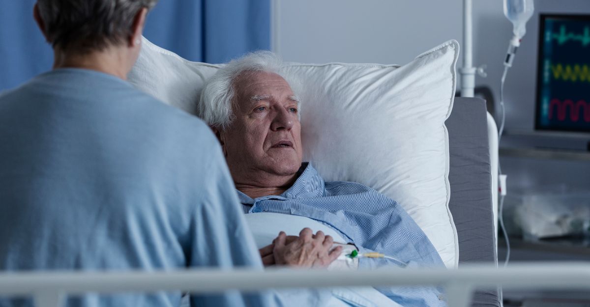 Uomo anziano a letto in ospedale con una persona al suo fianco.