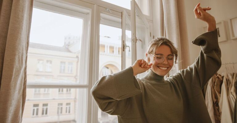 Una giovane donna dalla pelle chiara e divertente balla da sola a casa durante il giorno, con gli occhi chiusi. Una bionda con i capelli raccolti indossa occhiali trasparenti e un maglione oversize.
