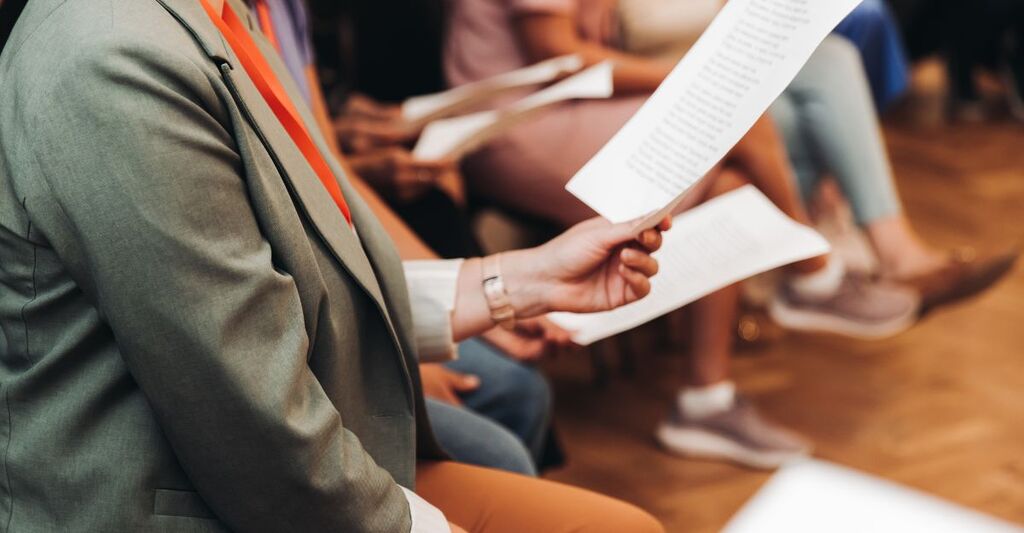 Prova del coro, lezione di canto nella sala dell'auditorium, gruppo sul palco tengono in mano dei fogli e cantano. Lezione di canto d'orchestra in una scuola di musica con un insegnante, adulti cantano in coro.