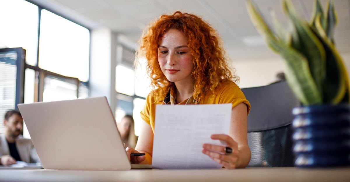 Giovane donna dai capelli rossi naturali guarda un foglio mentre è seduta alla scrivani in ufficio