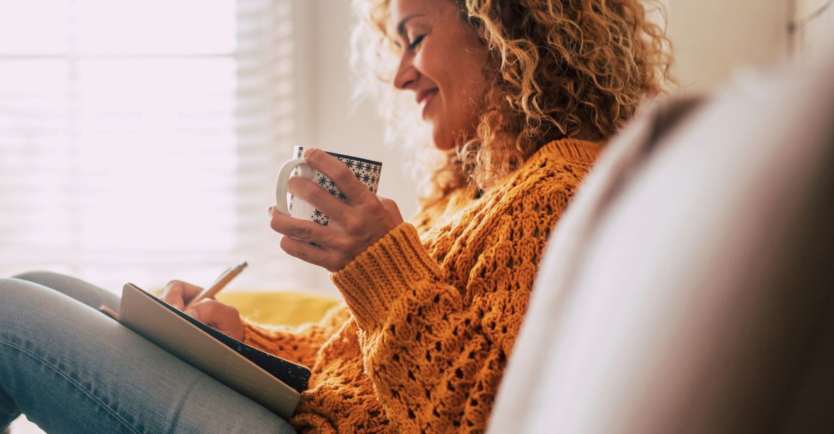 Una donna carina e felice a casa scrive appunti su un diario mentre beve una tazza di tè, si riposa e si rilassa prendendosi una pausa. Colori autunnali.