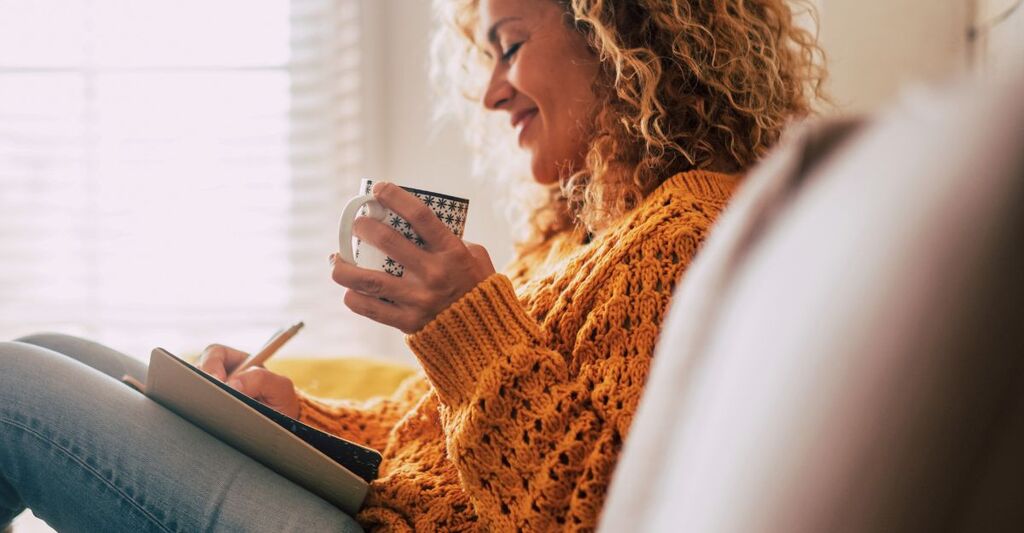 Una donna carina e felice a casa scrive appunti su un diario mentre beve una tazza di tè, si riposa e si rilassa prendendosi una pausa. Colori autunnali.