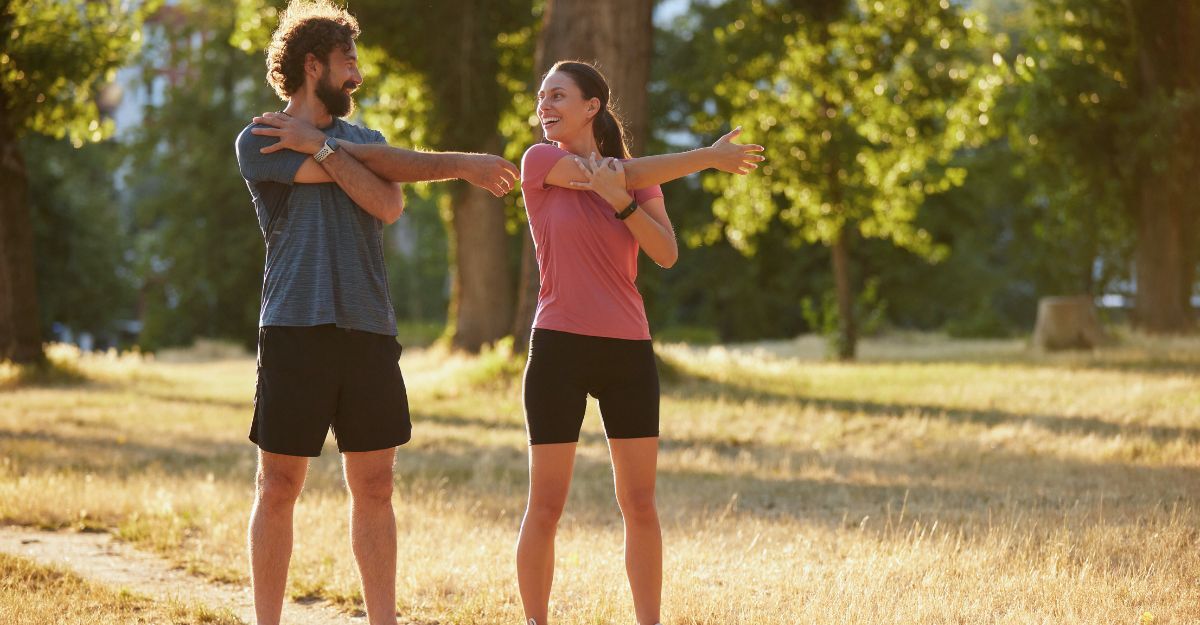 Due persone eseguono esercizi di stretching dinamico in un parco soleggiato, promuovendo la salute e il benessere attraverso l'attività fisica e il contatto con la natura.