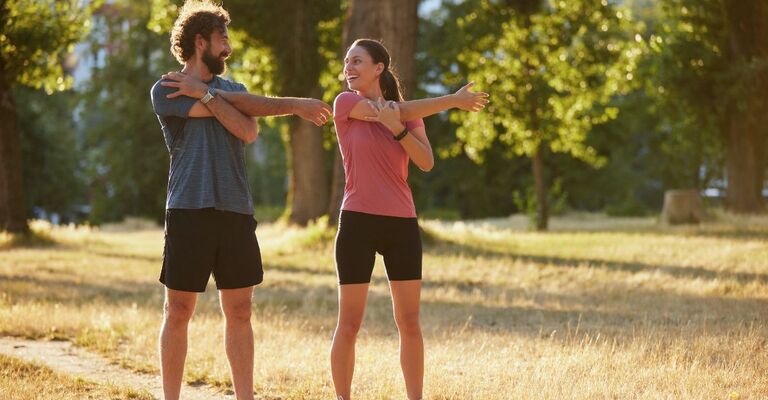 Due persone eseguono esercizi di stretching dinamico in un parco soleggiato, promuovendo la salute e il benessere attraverso l'attività fisica e il contatto con la natura.