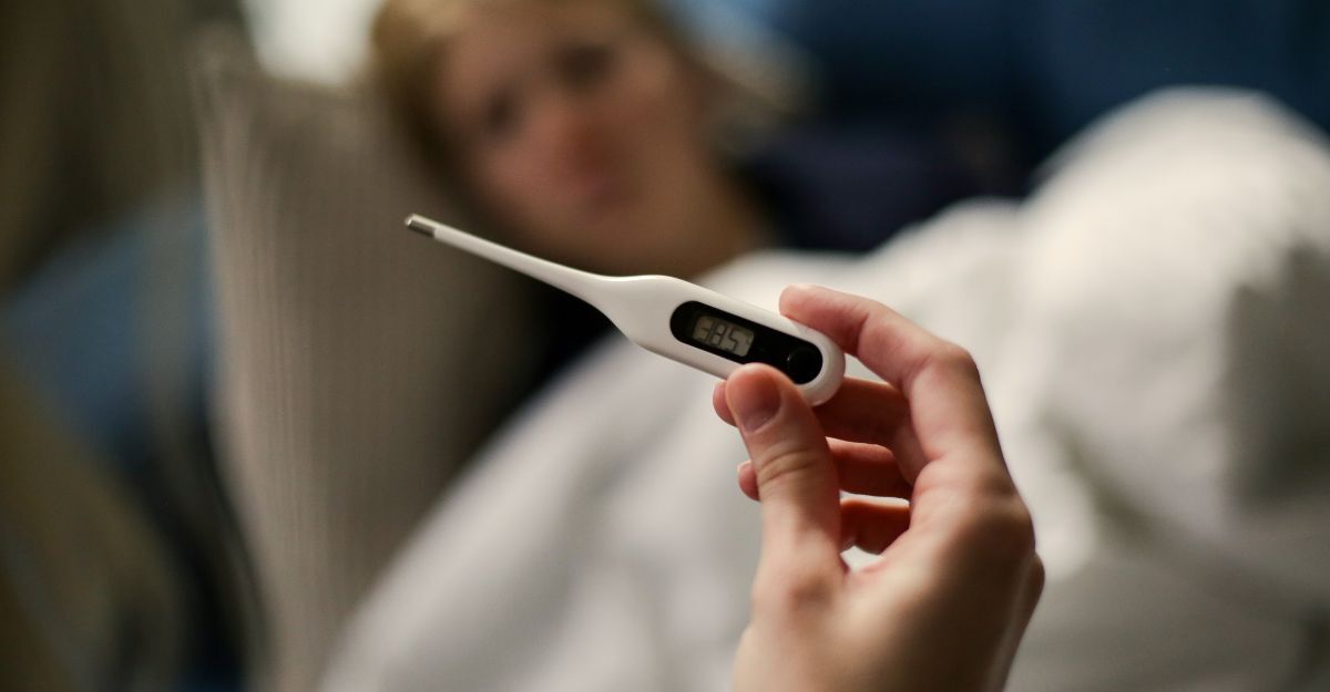 Una mano tiene in primo piano un termometro digitale che segna 38,5°C, con una persona sfocata a letto sullo sfondo