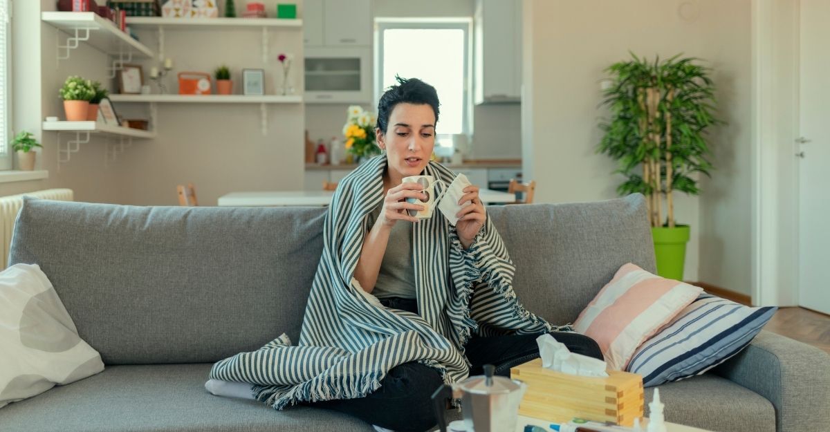 Una donna seduta sul divano, avvolta in una coperta, mentre tiene una tazza calda e un fazzoletto