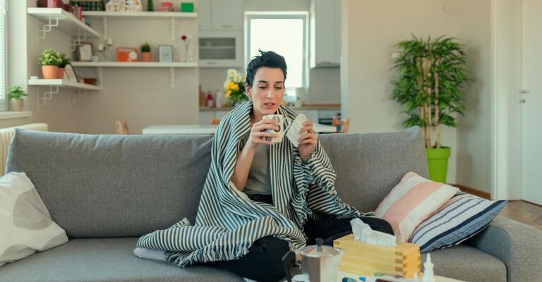 Una donna seduta sul divano, avvolta in una coperta, mentre tiene una tazza calda e un fazzoletto