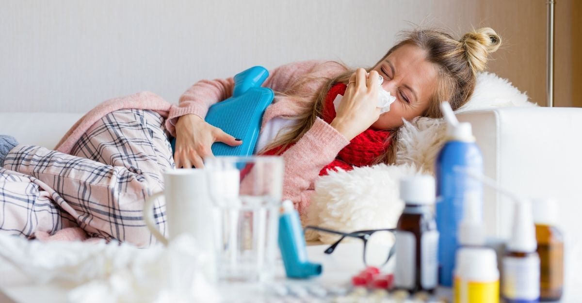 Una donna distesa sul divano che soffia il naso, con una borsa dell'acqua calda e diversi medicinali in primo piano