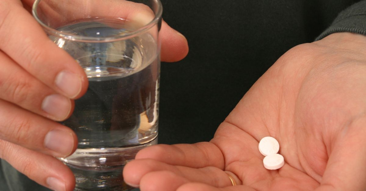 Una persona tiene due piccole pillole bianche sul palmo della mano e un bicchiere d'acqua nell'altra