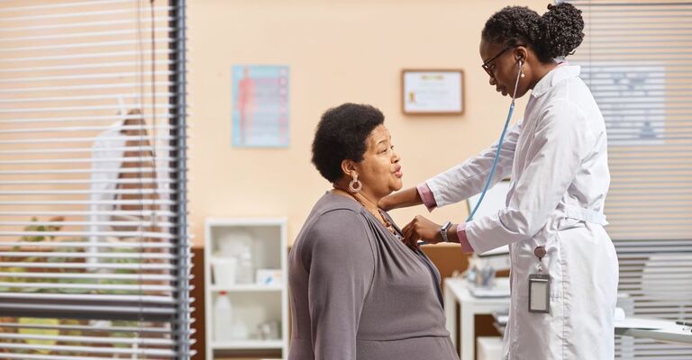 Vista laterale di una donna anziana di colore seduta sul lettino da terapia in uno studio medico che espira mentre un'operatrice sanitaria ascolta i suoni polmonari