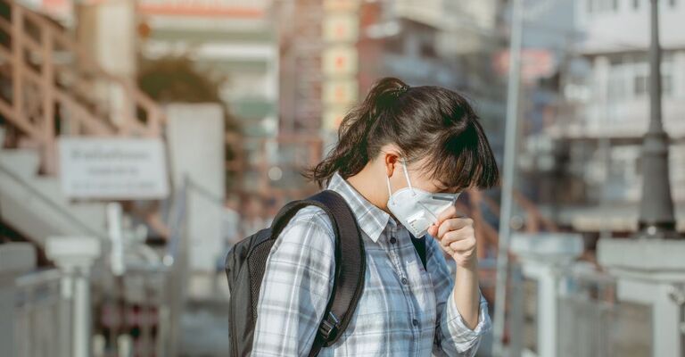 Una ragazza con zaino e mascherina filtrante tossisce mentre cammina in una zona urbana offuscata dallo smog