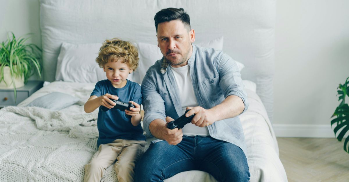 Un uomo e un bambino seduti sul bordo di un letto mentre giocano concentrati a un videogioco con i controller
