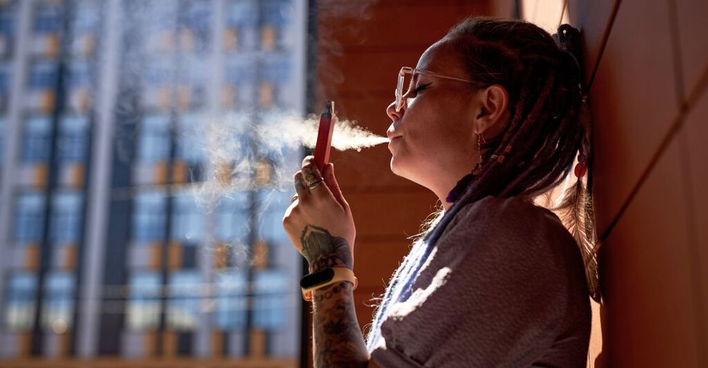Giovane donna con una sigaretta elettronica in mano in piedi vicino al muro di un edificio che rilascia una nuvola di vapore dalla bocca mentre fuma