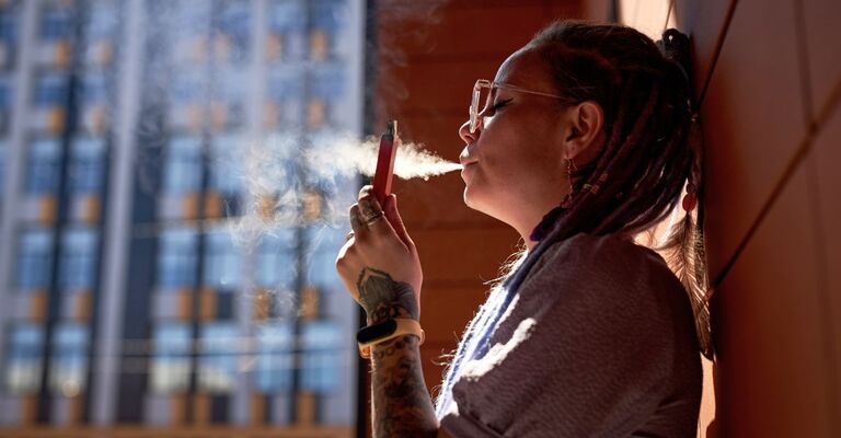 Giovane donna con una sigaretta elettronica in mano in piedi vicino al muro di un edificio che rilascia una nuvola di vapore dalla bocca mentre fuma