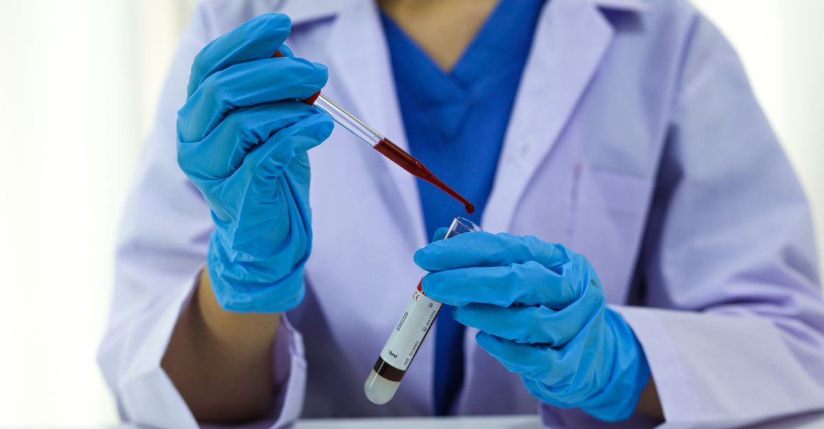 Un ricercatore in camice blu e mascherina maneggia delle provette in un laboratorio accanto a un microscopio ottico.