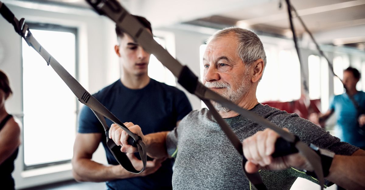 Un gruppo di anziani in palestra con un giovane allenatore che ne aiuta uno a fare esercizi con il TRX