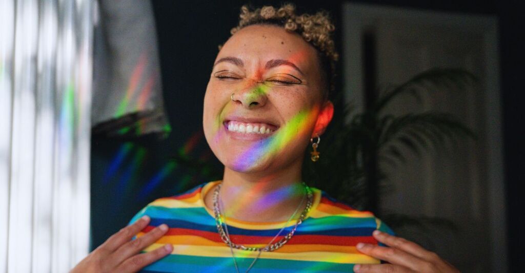 Ragazza in primo piano che sorride con gli occhi chiusi mentre la luce del sole scomposta in colori le arriva in faccia