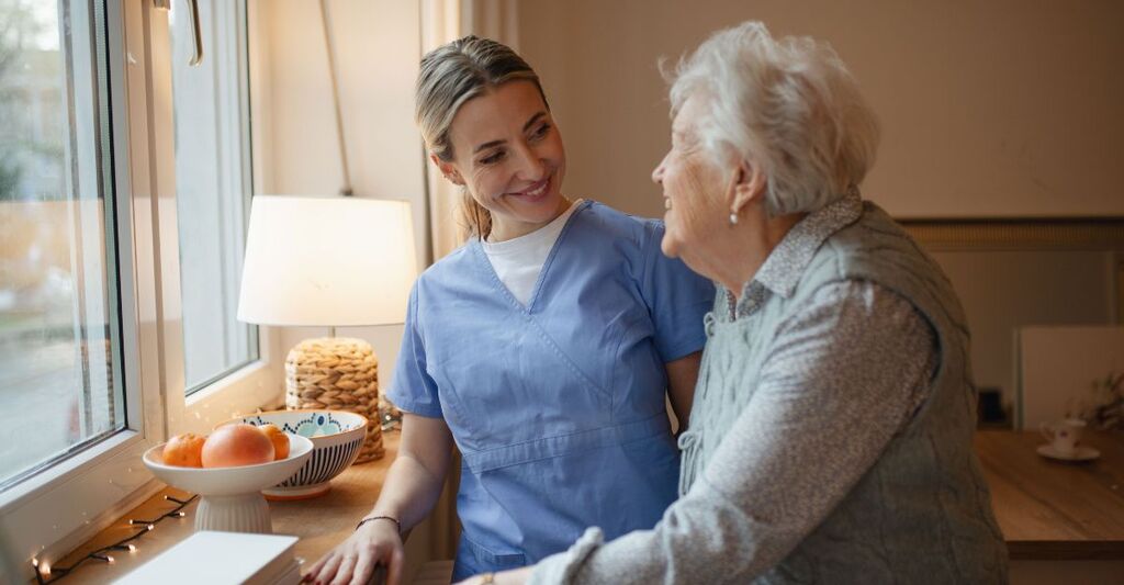 Una giovane infermiera in divisa azzurra sorride a una donna anziana vicino a una finestra in un ambiente domestico accogliente