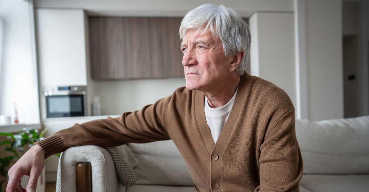 Primo piano di un uomo anziano con i capelli grigi e un cardigan marrone, seduto in soggiorno con un'espressione pensierosa