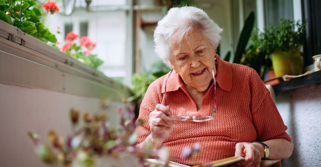 Una signora anziana a casa in veranda legge un libro.