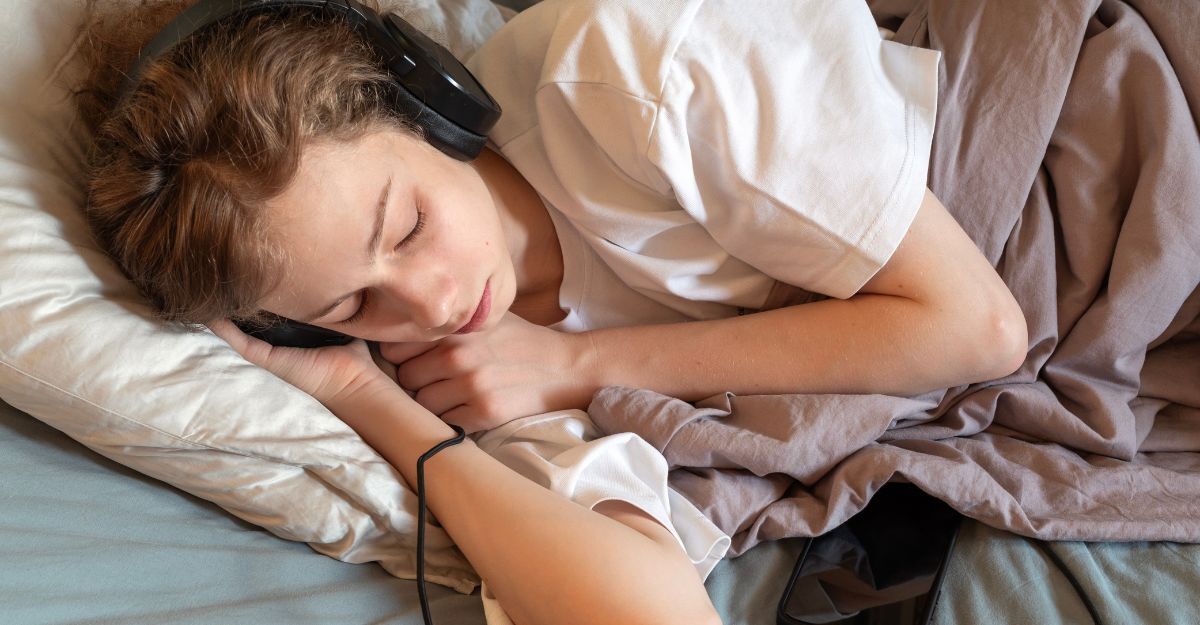 Una ragazza dorme nel letto indossando grandi cuffie nere collegate a uno smartphone