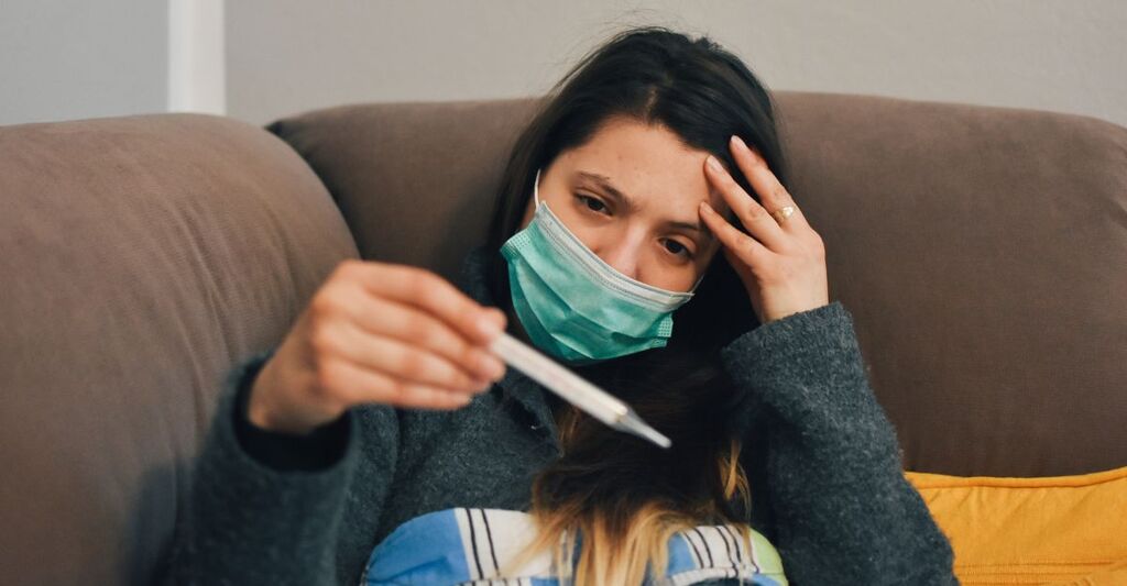 Una ragazza con la mascherina che si prova la febbre sul divano