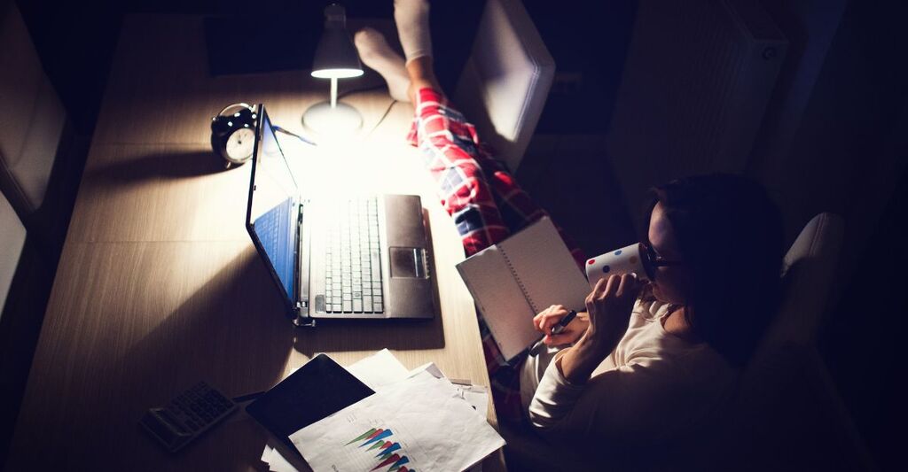 Una persona lavora al computer di notte con una lampada da tavolo, circondata da documenti e appunti