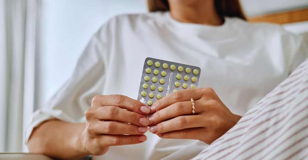 Giovane donna caucasica adulta che tiene in mano un blister di pillole anticoncezionali orali con entrambe le mani, seduta in un ambiente chiuso, con il viso parzialmente visibile, concentrata sulla confezione del farmaco