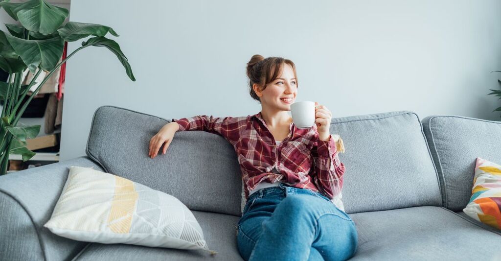 Giovane donna sorridente seduta sul divano che guarda altrove mentre beve un caffè o un tè. Giovane donna bruna che si rilassa dopo le pulizie domestiche. Ritratto di donna rilassata che riposa a casa.