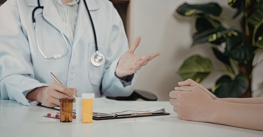 Primo piano delle mani di un medico che scrive su documenti medici mentre parla con un paziente; sul tavolo sono presenti flaconi di medicinali