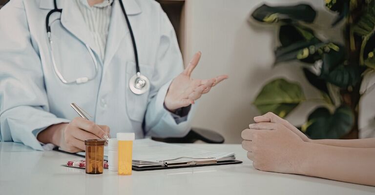 Primo piano delle mani di un medico che scrive su documenti medici mentre parla con un paziente; sul tavolo sono presenti flaconi di medicinali