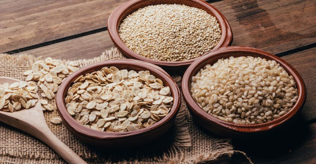 Tre ciotole di terracotta contenenti fiocchi d'avena, quinoa e riso integrale su un tavolo di legno