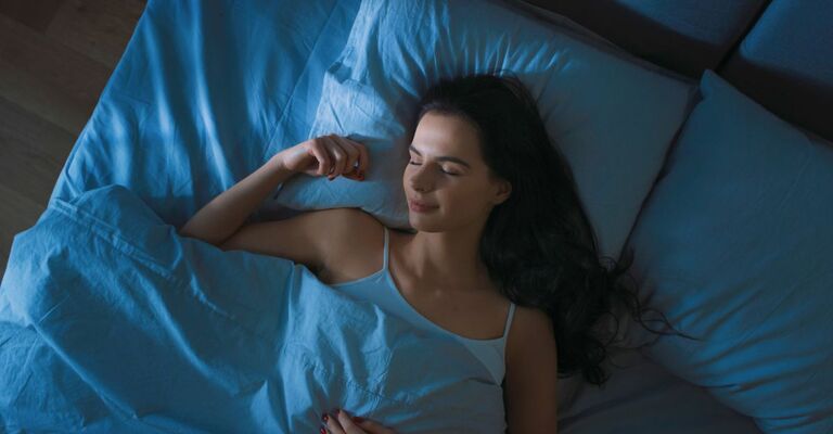 Vista dall'alto di una bellissima giovane donna che dorme comodamente su un letto nella sua camera da letto di notte. Colori notturni blu con la luce fredda e debole del lampione che filtra dalla finestra.
