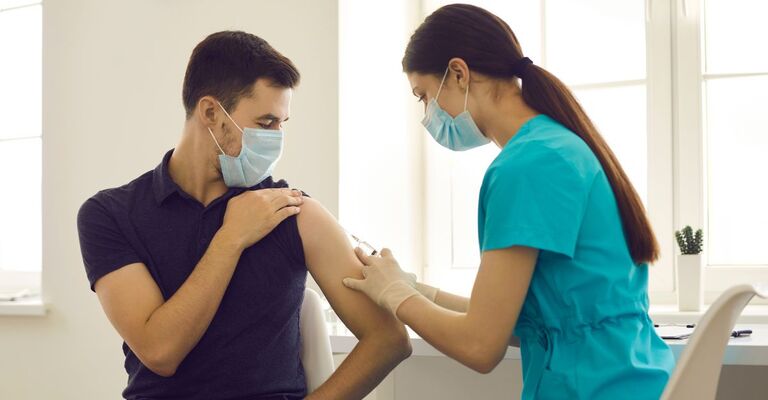 Un'infermiera in divisa azzurra e mascherina inietta un vaccino sulla spalla di un uomo seduto