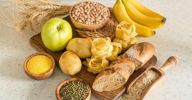 Selezione di carboidrati complessi: pane integrale, pasta, patate, legumi, riso, cereali e frutta su un tagliere in legno