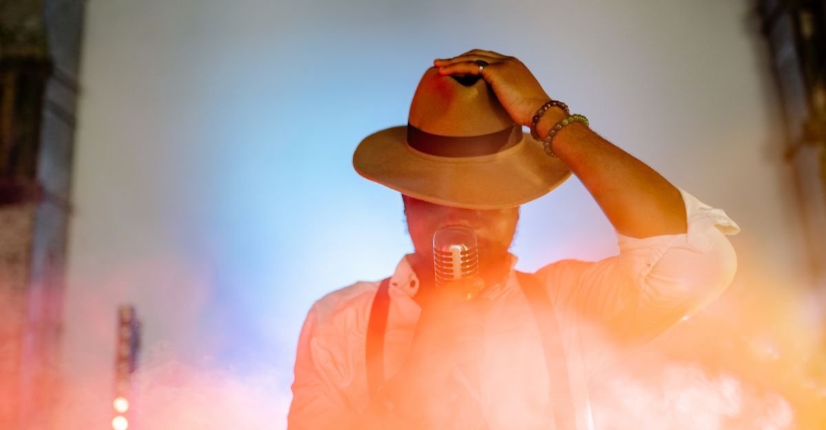 Un cantante con cappello e bretelle che tiene un microfono vintage, immerso in una nebbia colorata dalle luci di scena