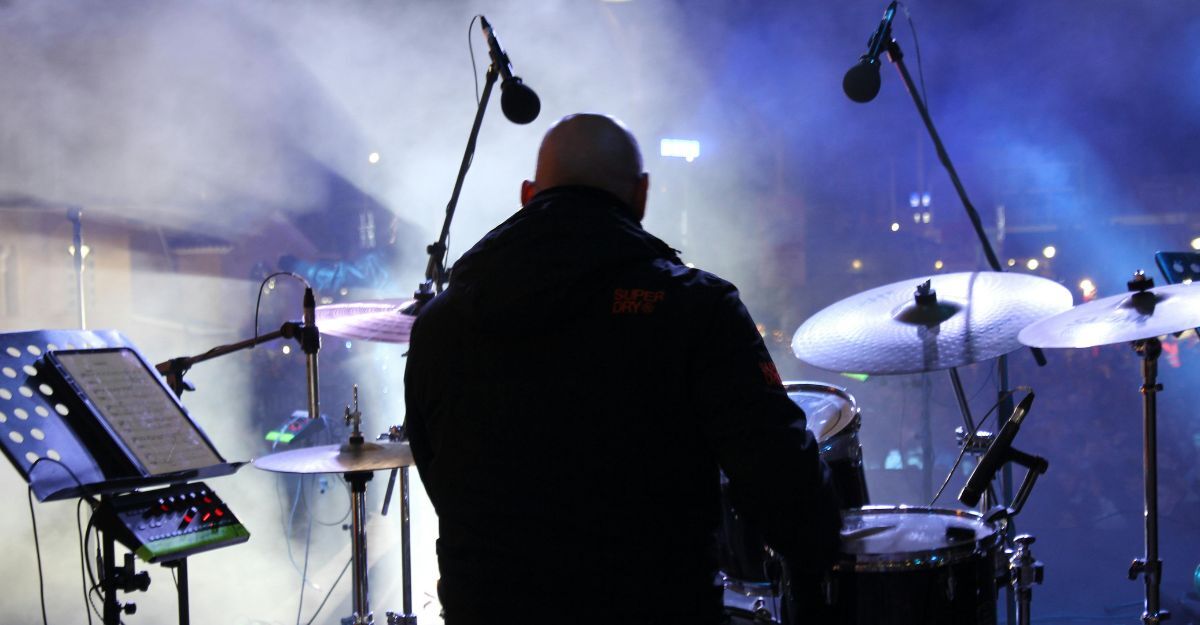 Un batterista di spalle sul palco, circondato da fumo scenico e luci blu durante un concerto dal vivo