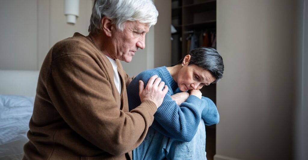 Un uomo anziano poggia con empatia una mano sulla spalla di una donna seduta a terra, che appare profondamente triste e assorta nei suoi pensieri