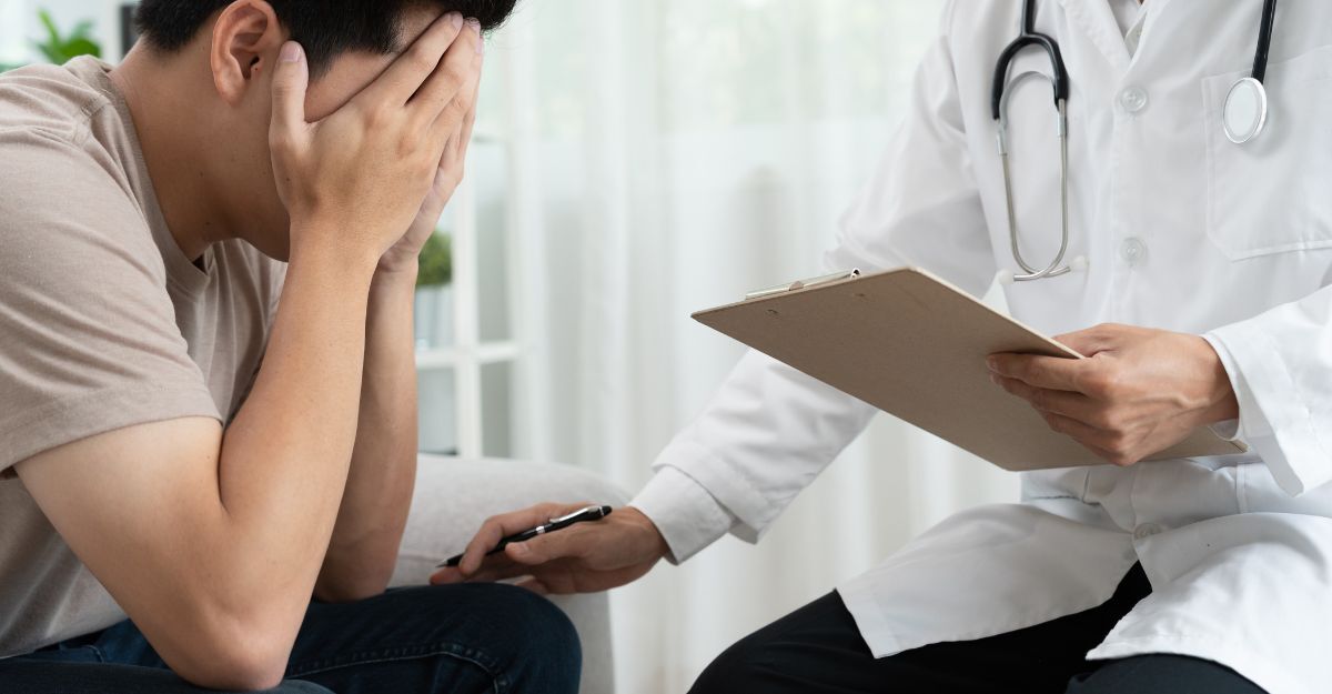Un medico in camice bianco conforta un giovane uomo visibilmente scosso che si copre il volto con le mani durante un colloquio clinico