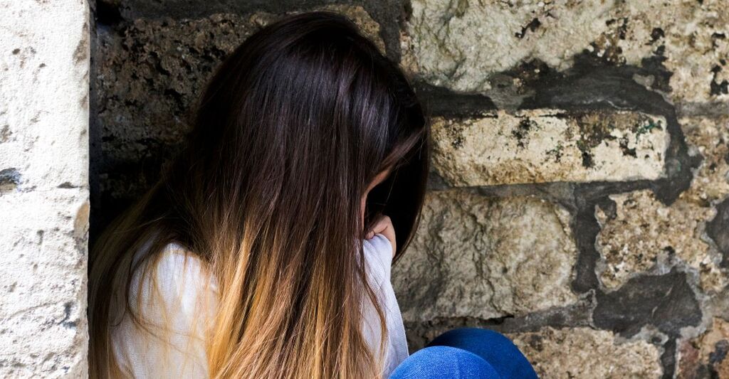 Una ragazza con i capelli lunghi rannicchiata contro un muro di pietra, con il volto nascosto, in segno di tristezza