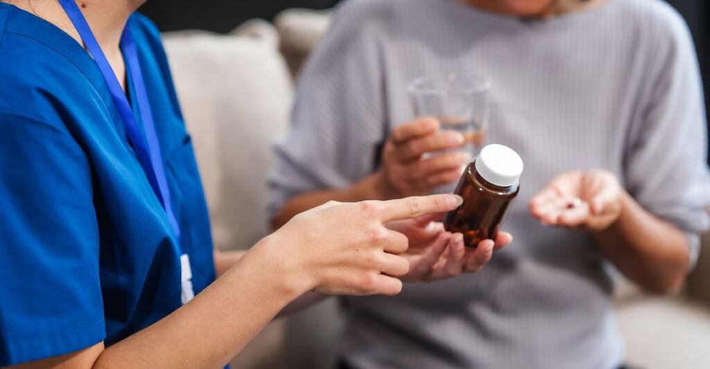 Una dottoressa caucasica offre un bicchiere d'acqua e delle pillole a un anziano paziente asiatico mentre sono seduti insieme sul divano